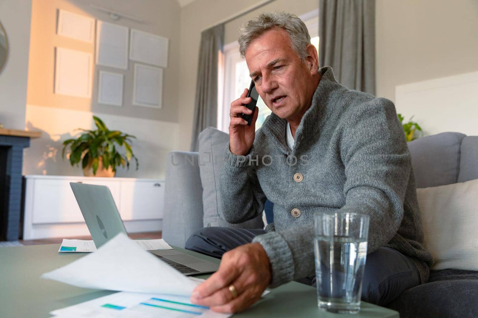 Focused caucasian senior man sitting on sofa, doing paperwork, making call and using laptop. active retirement lifestyle at home.