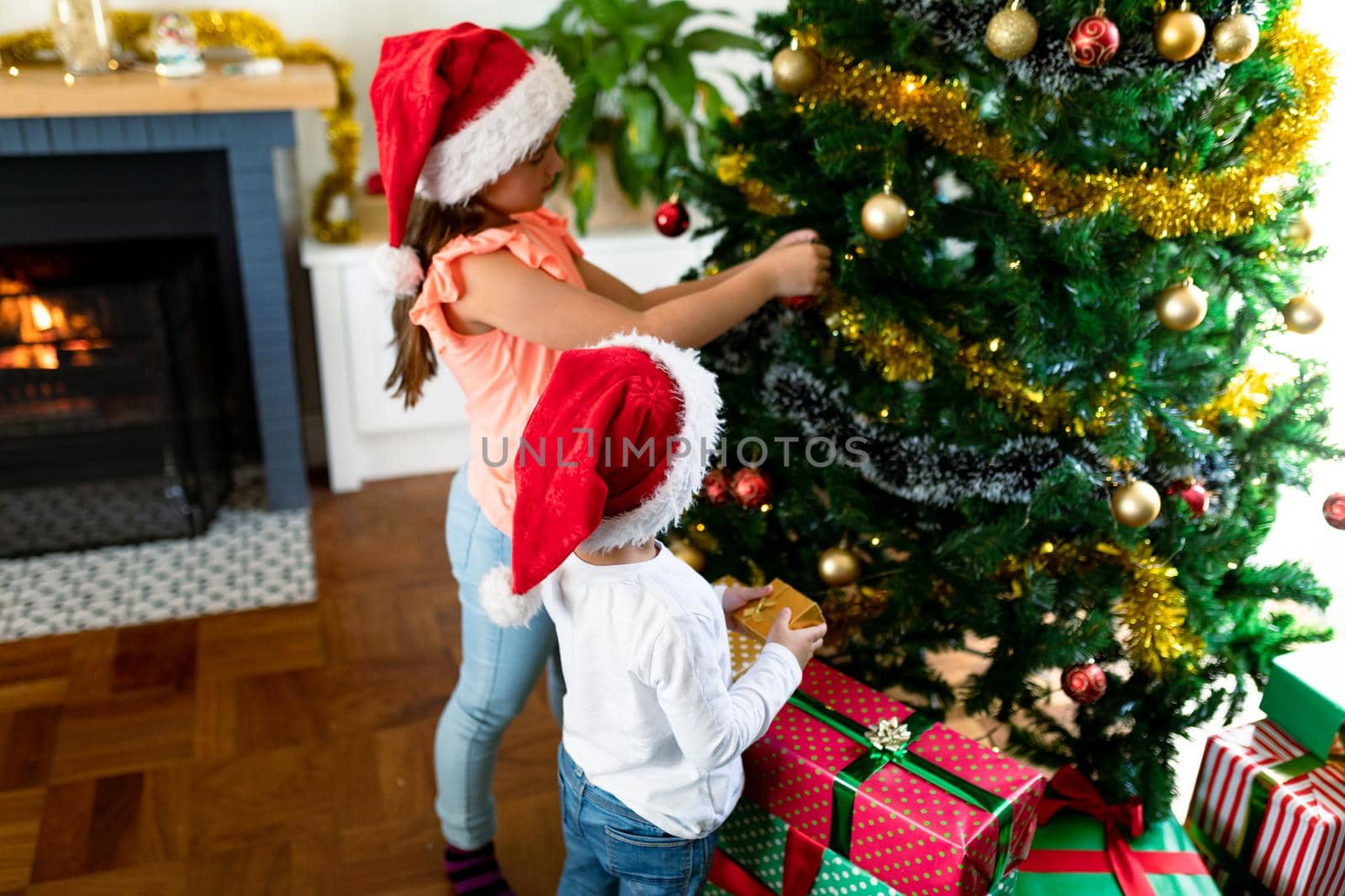 Happy caucasian sibling wearing santa hats, decorating christmas tree. family christmas time and festivity together at home.