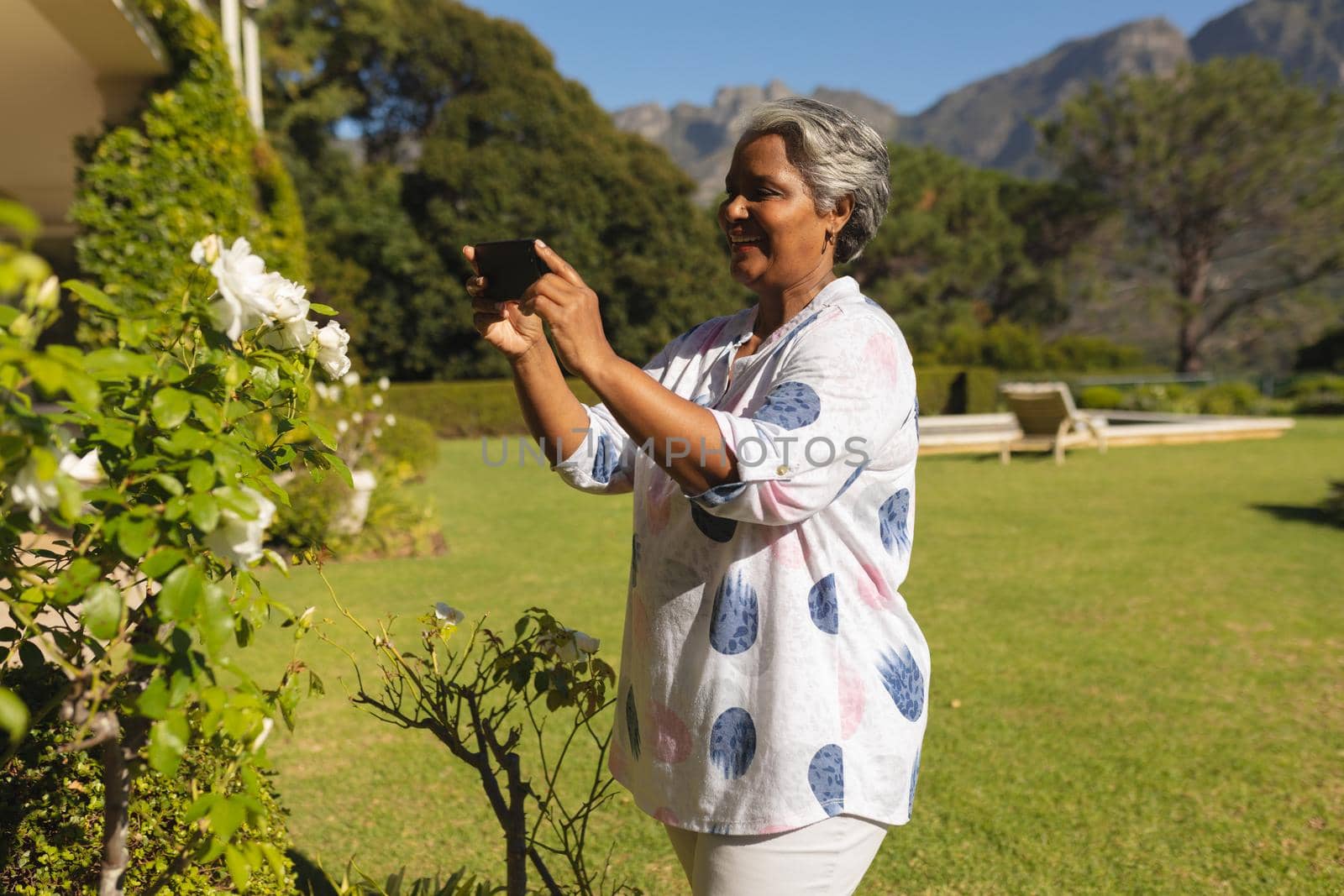 Senior african american woman taking photos with smartphone in sunny garden. retreat, retirement and happy senior lifestyle concept.