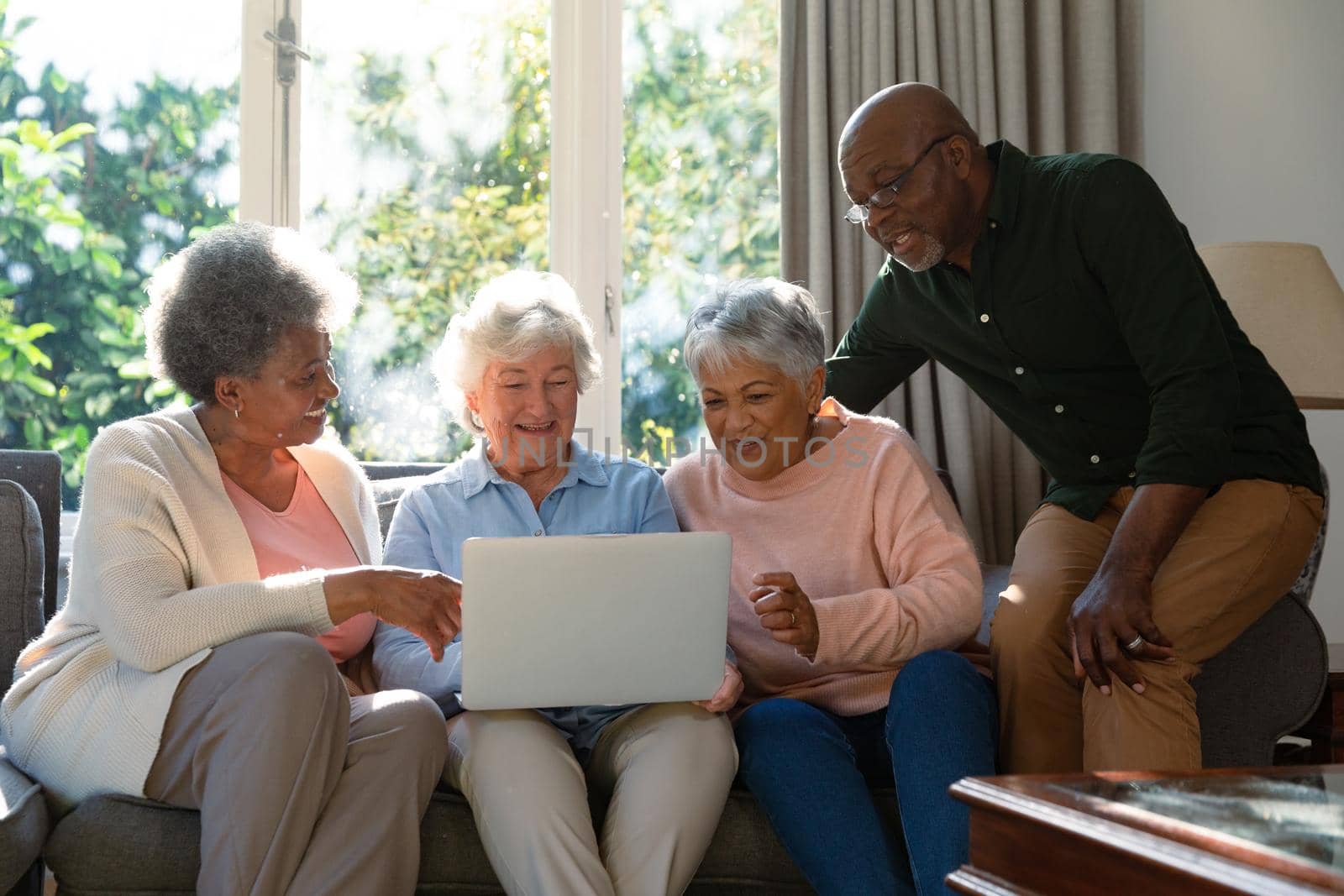 Three happy diverse senior woman and african american male friend sitting on sofa and using laptop. retirement lifestyle relaxing at home with technology.