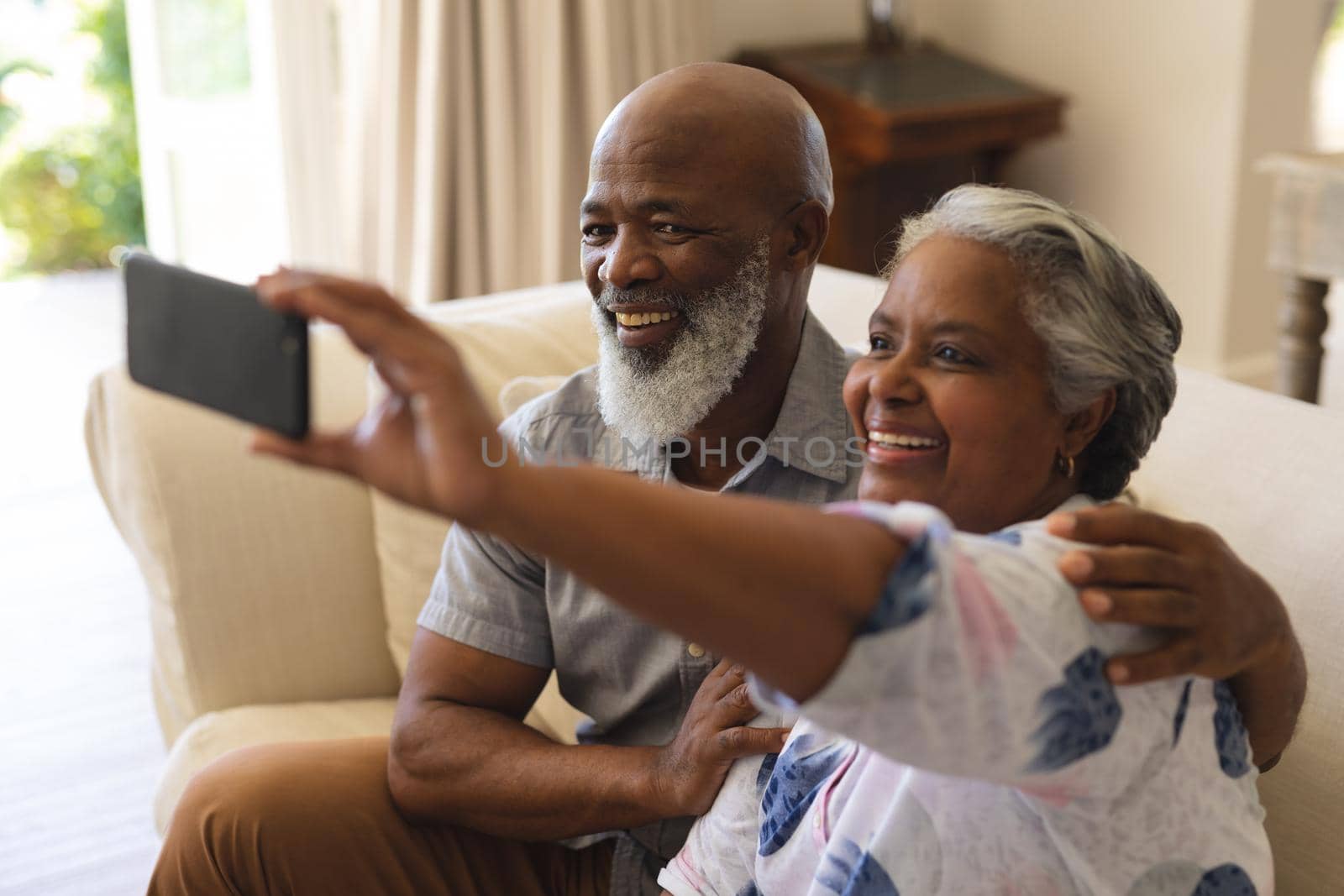 Senior african american couple sitting on sofa using smartphone taking selfies. retreat, retirement and happy senior lifestyle concept.