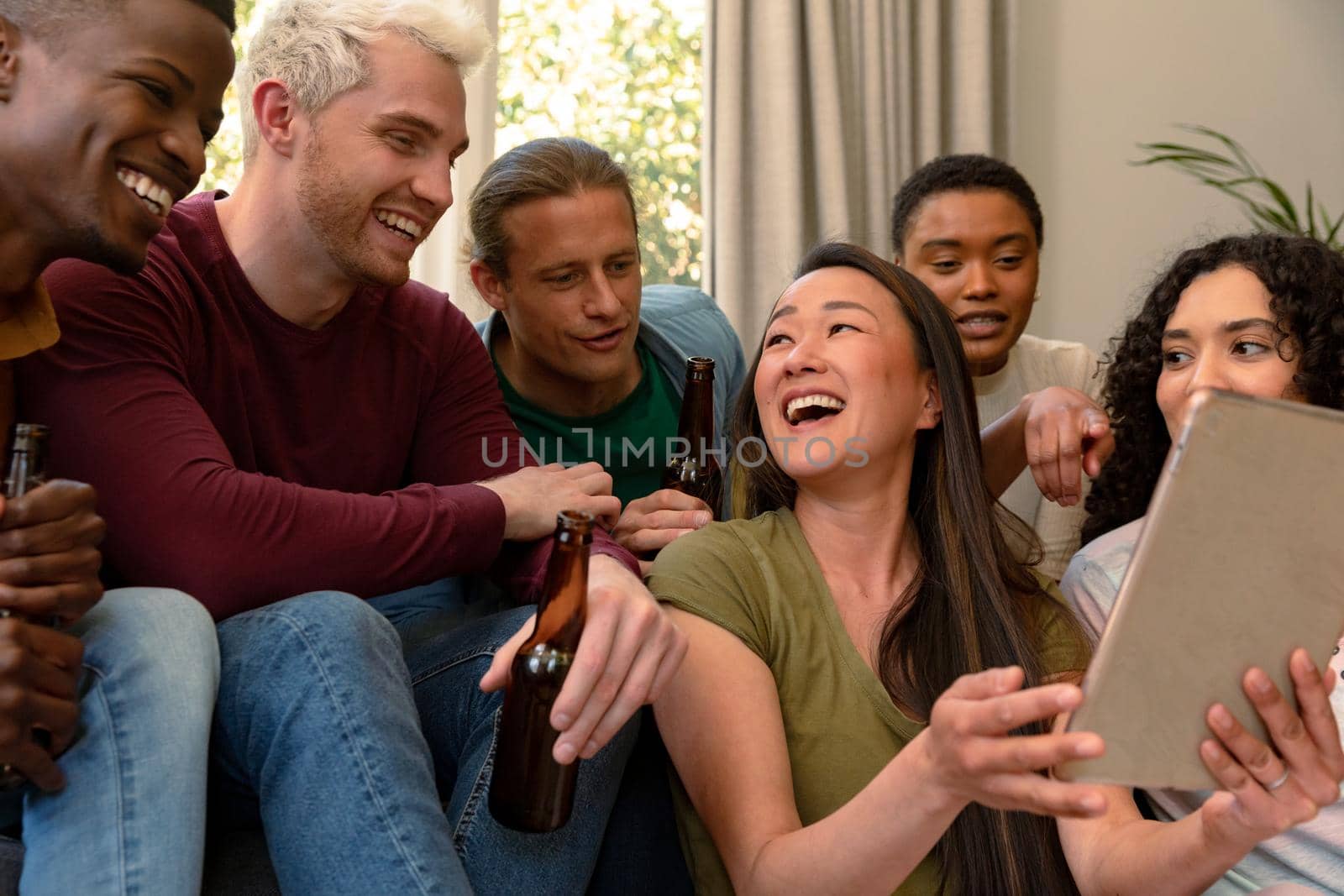 Group of happy diverse female and male friends drinking beer together and using tablet. socialising with friends at home.