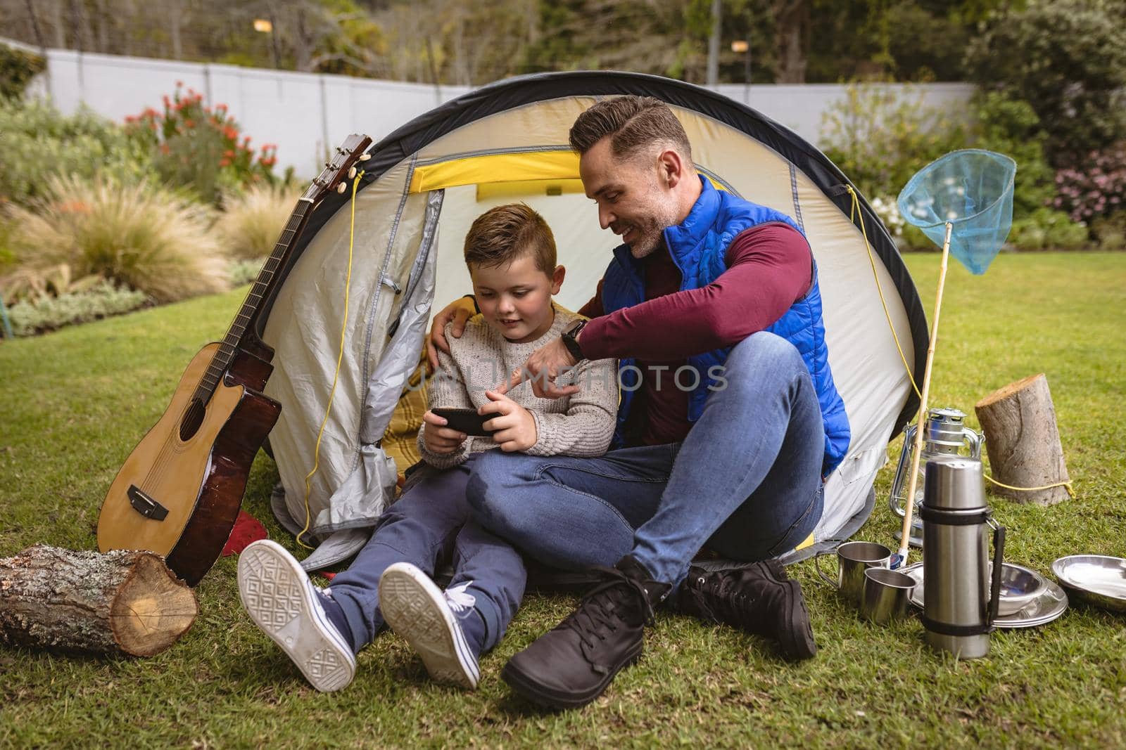 Caucasian father and son using smartphone while sitting in a tent in the garden. fatherhood and love concept
