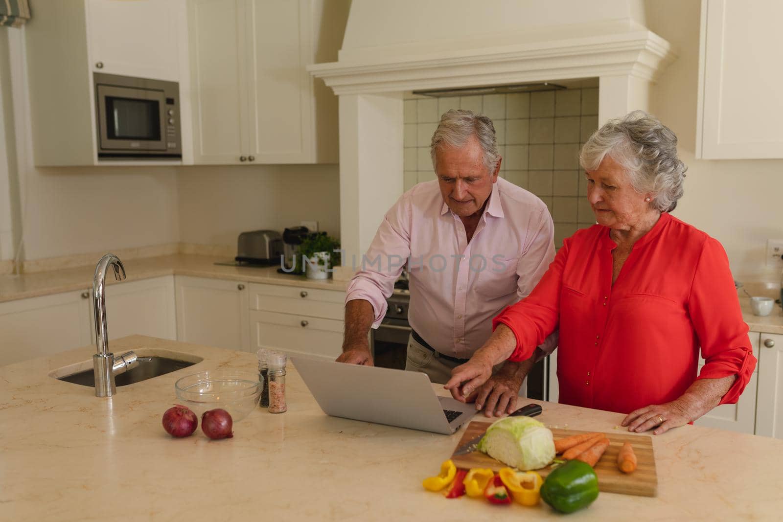 Senior caucasian couple cooking together and using laptop in kitchen. retreat, retirement and happy senior lifestyle concept.