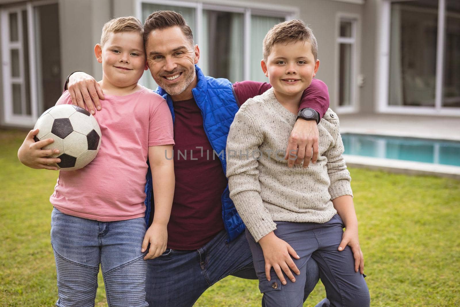 Portrait of caucasian father and two sons smiling while holding football in the garden. fatherhood and love concept