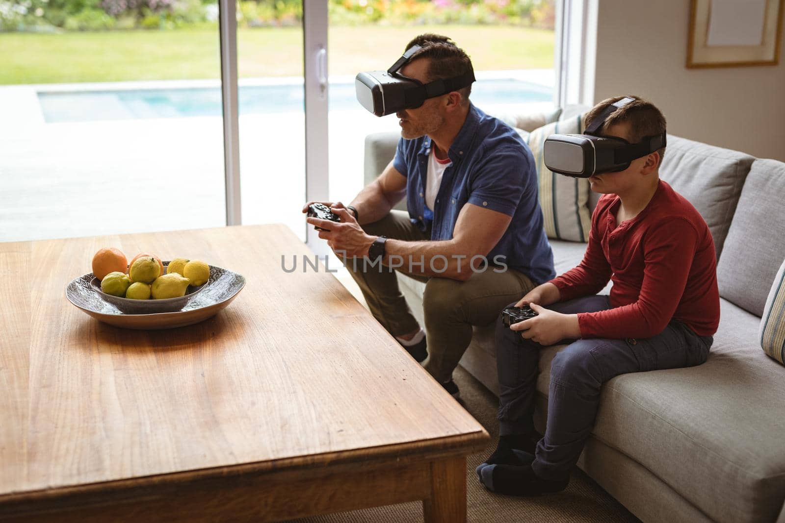 Caucasian father and son wearing vr headsets playing video games sitting on the couch at home by Wavebreakmedia