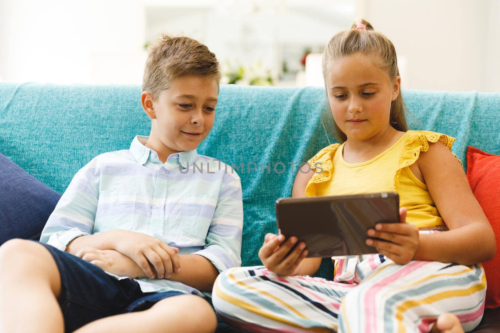 Happy caucasian brother and sister sitting on couch and using tablet in living room. childhood leisure time, fun and discovery at at home using technology.