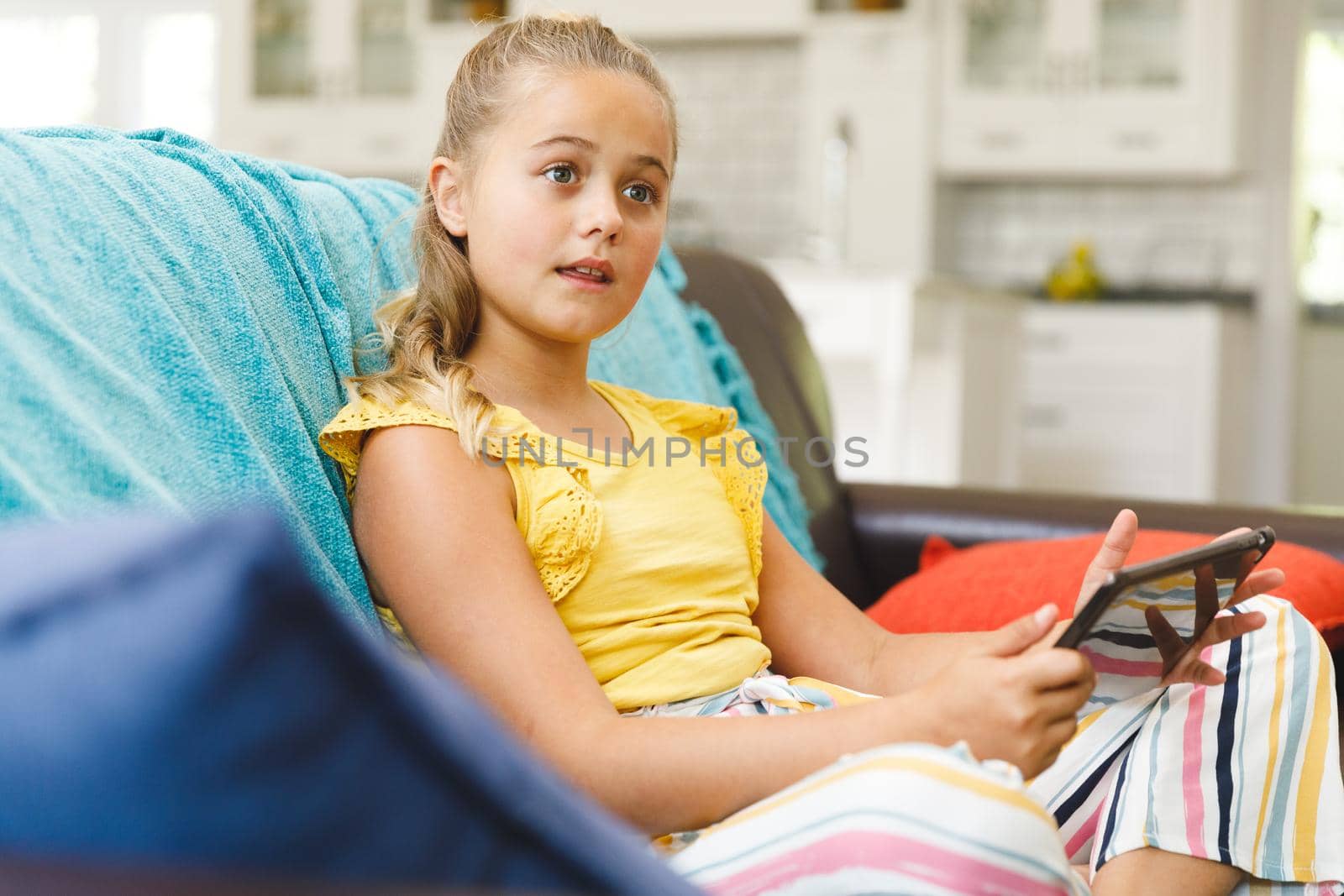 Caucasian girl sitting on couch and using tablet in living room. childhood leisure time, fun and discovery at at home using technology.