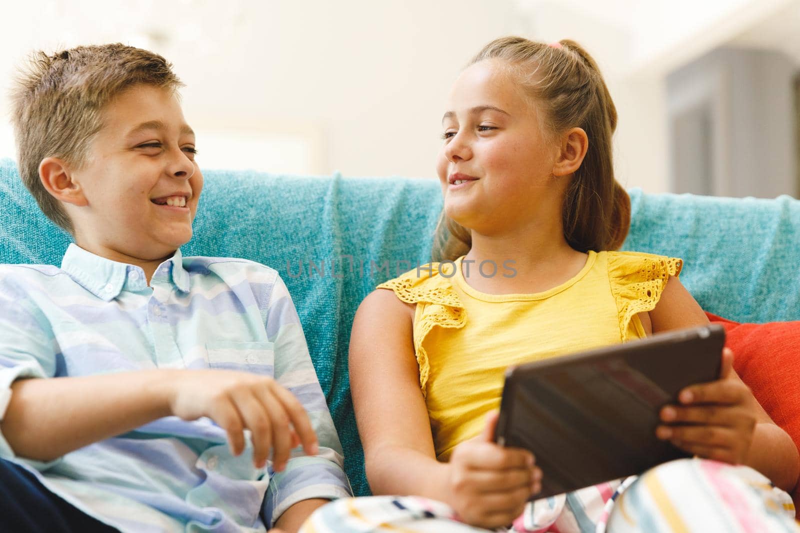 Happy caucasian brother and sister sitting on couch and using tablet in living room. childhood leisure time, fun and discovery at at home using technology.