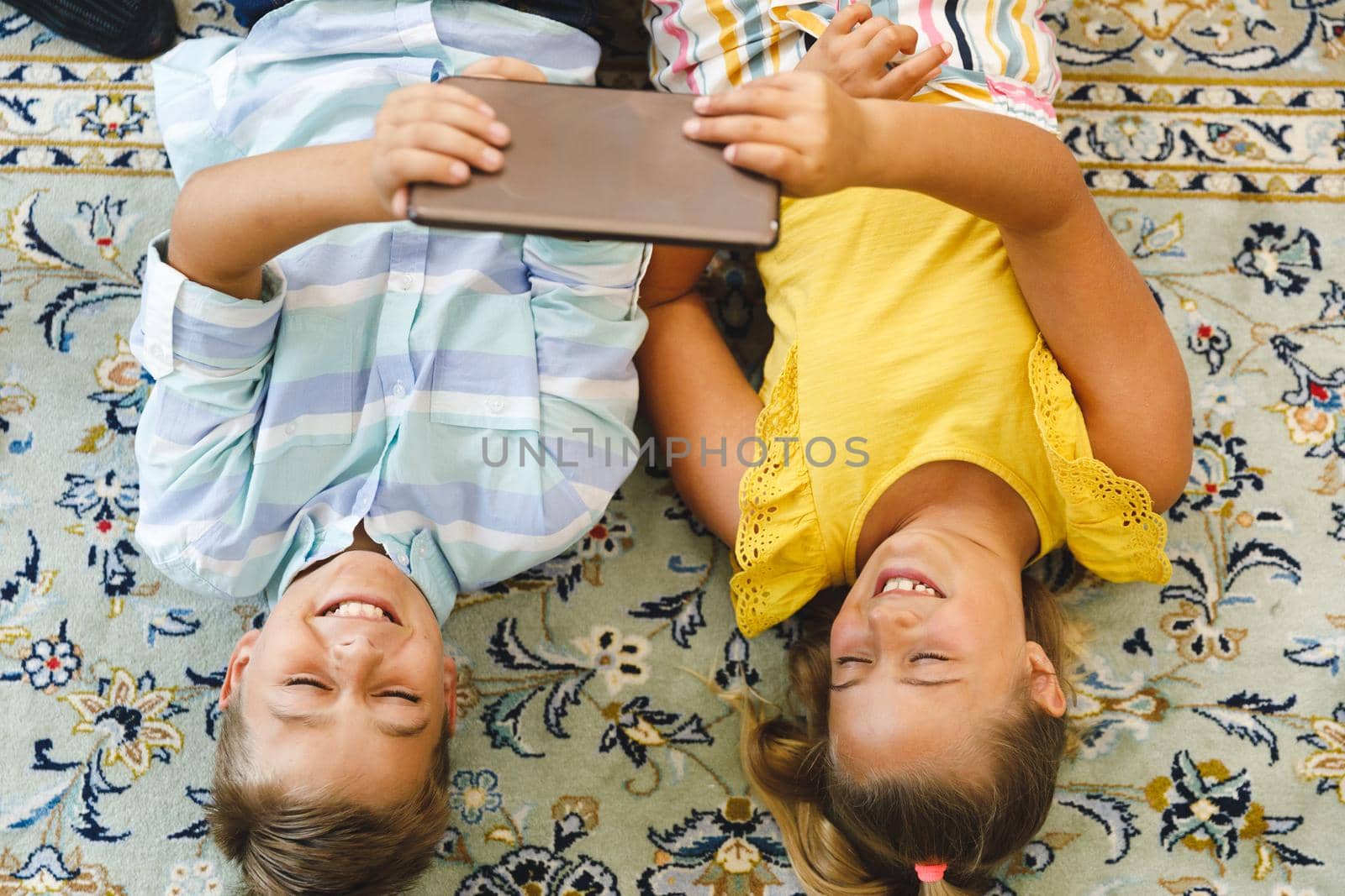 Smiling caucasian brother and sister lying in living room and using tablet. childhood leisure time, fun and discovery at at home using technology.