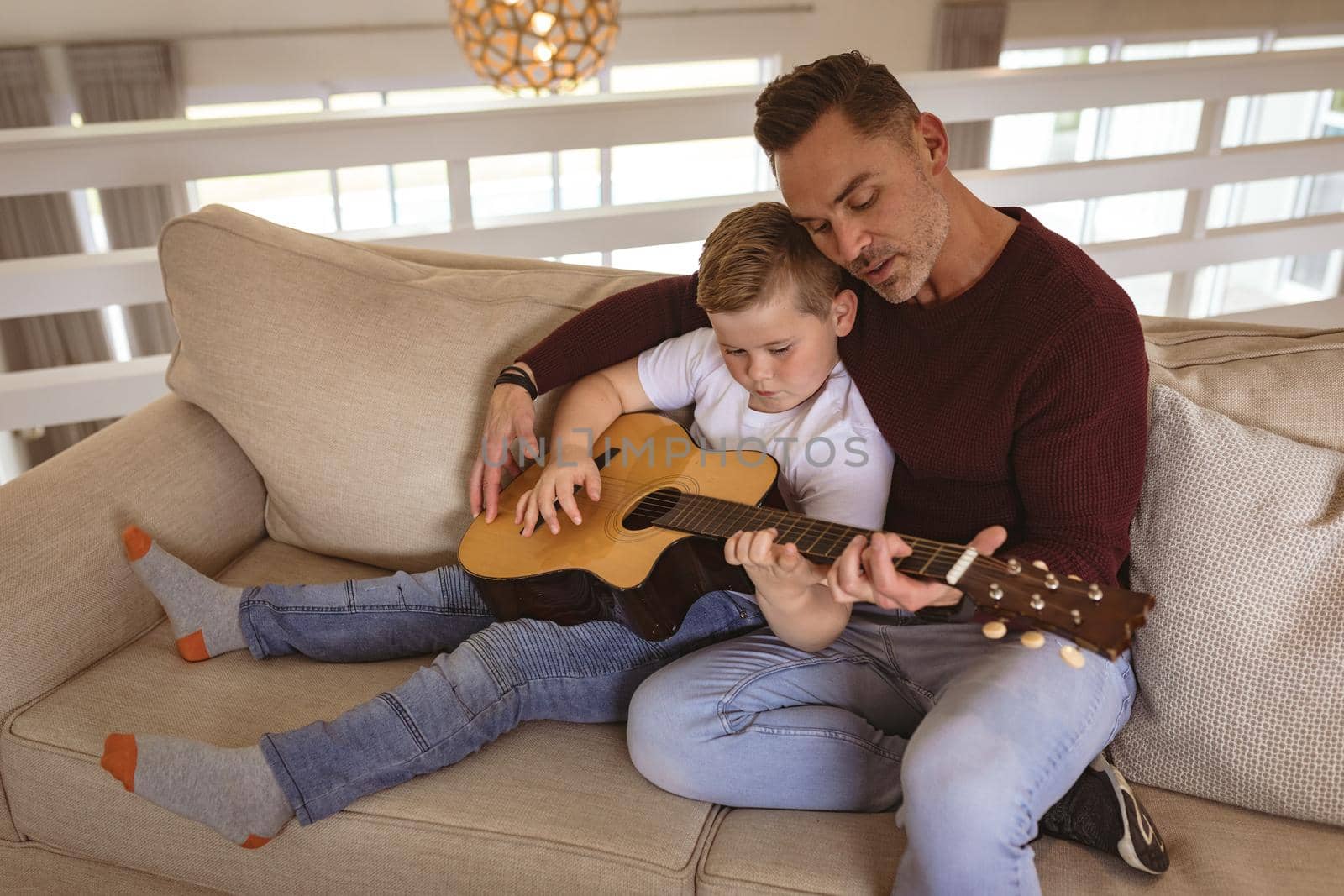 Caucasian father teaching his son to play guitar sitting on the couch at home. childhood and hobby concept