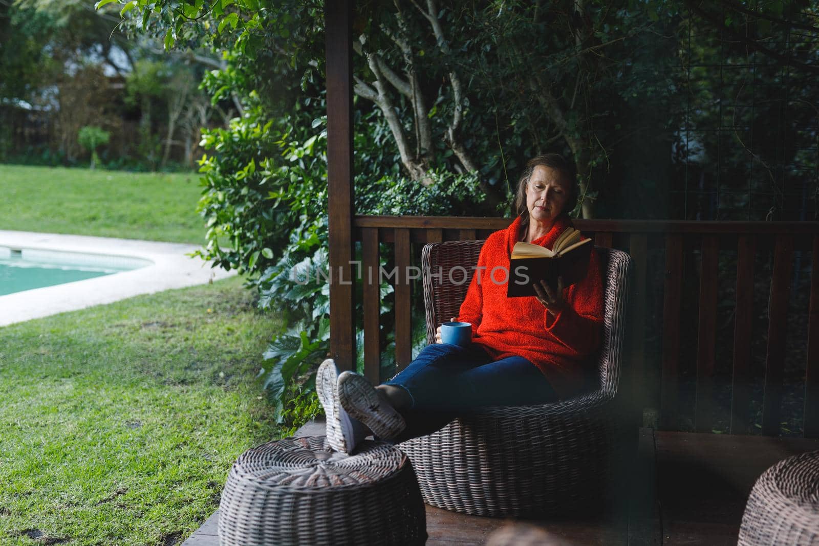 Happy senior caucasian woman sitting on chair, reading book and drinking coffee in garden by Wavebreakmedia