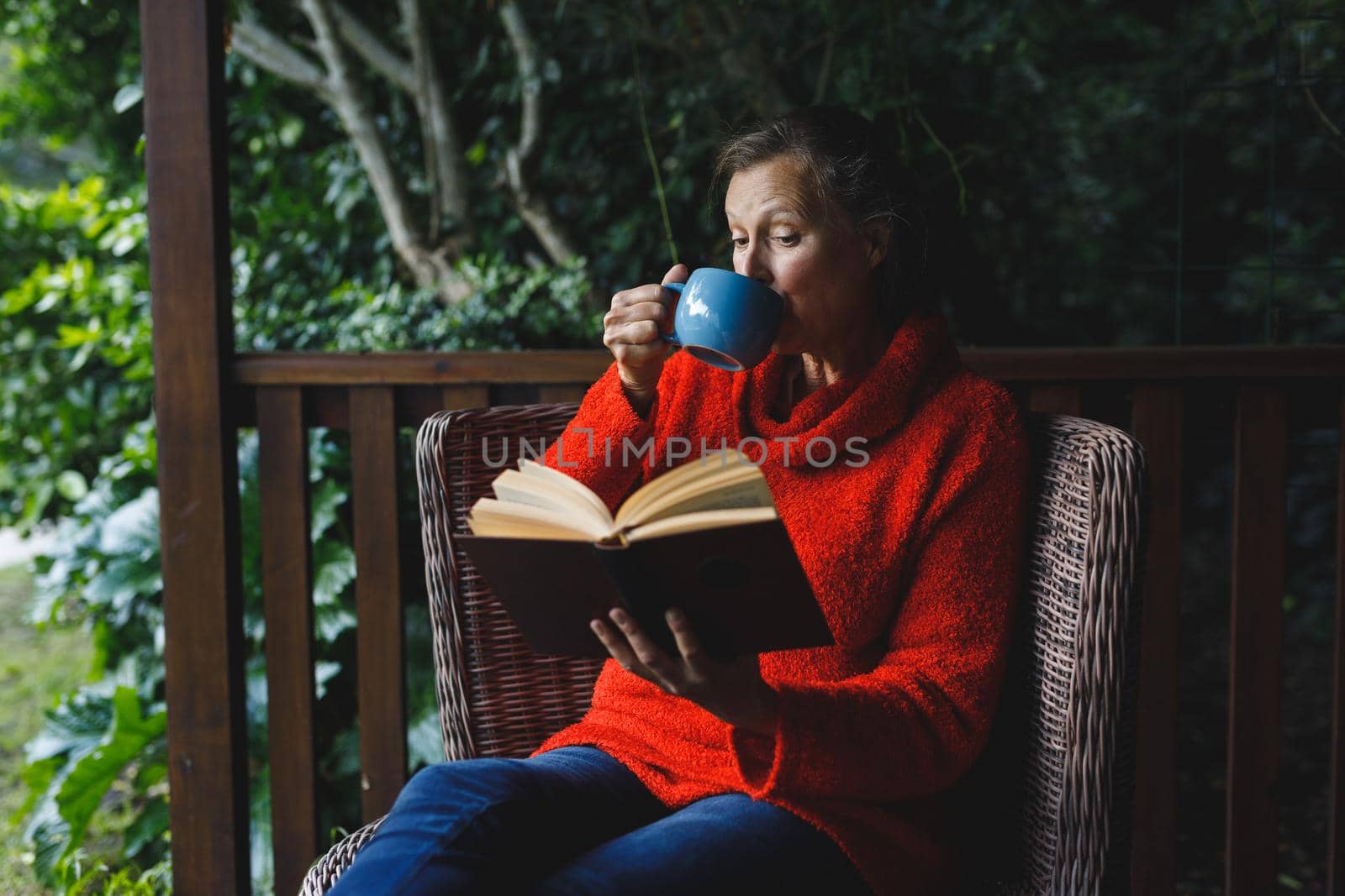 Happy senior caucasian woman sitting on chair, reading book and drinking coffee in garden by Wavebreakmedia