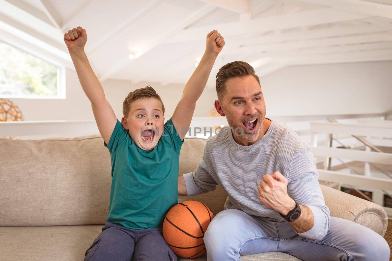 Caucasian father and son cheering while watching sports on television sitting on the couch at home by Wavebreakmedia