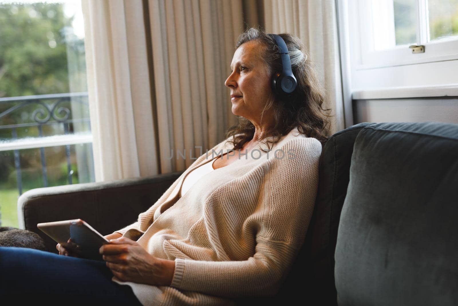 Happy senior caucasian woman in living room sitting on sofa, wearing headphones, using tablet by Wavebreakmedia