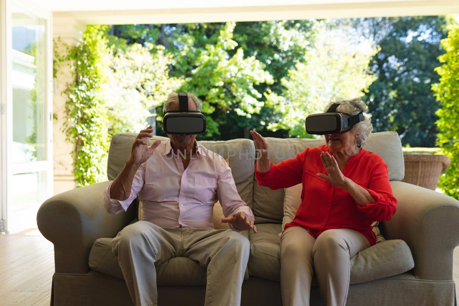 Senior caucasian couple sitting on sofa together wearing vr headset touching virtual screen. retreat, retirement and happy senior lifestyle concept.