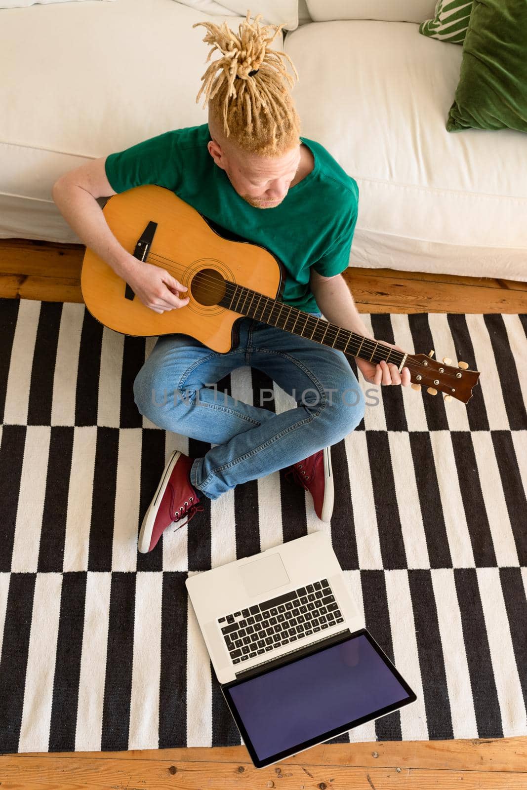 Albino african american man in the living room playing guitar and using laptop. leisure time using technology, relaxing at home.