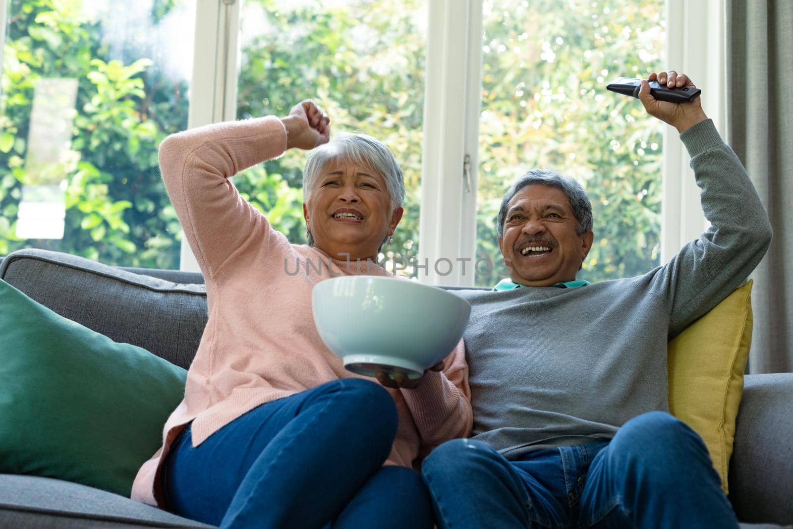 Two diverse senior couple sitting on sofa, watching tv and having fun. retirement lifestyle relaxing at home with technology.