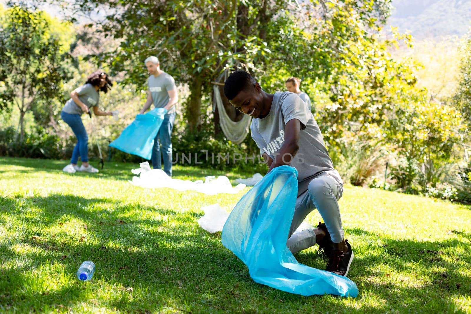Smiling african american man holding refuse sack, collecting plastic waste in field with friends. eco conservation volunteers doing countryside clean-up.