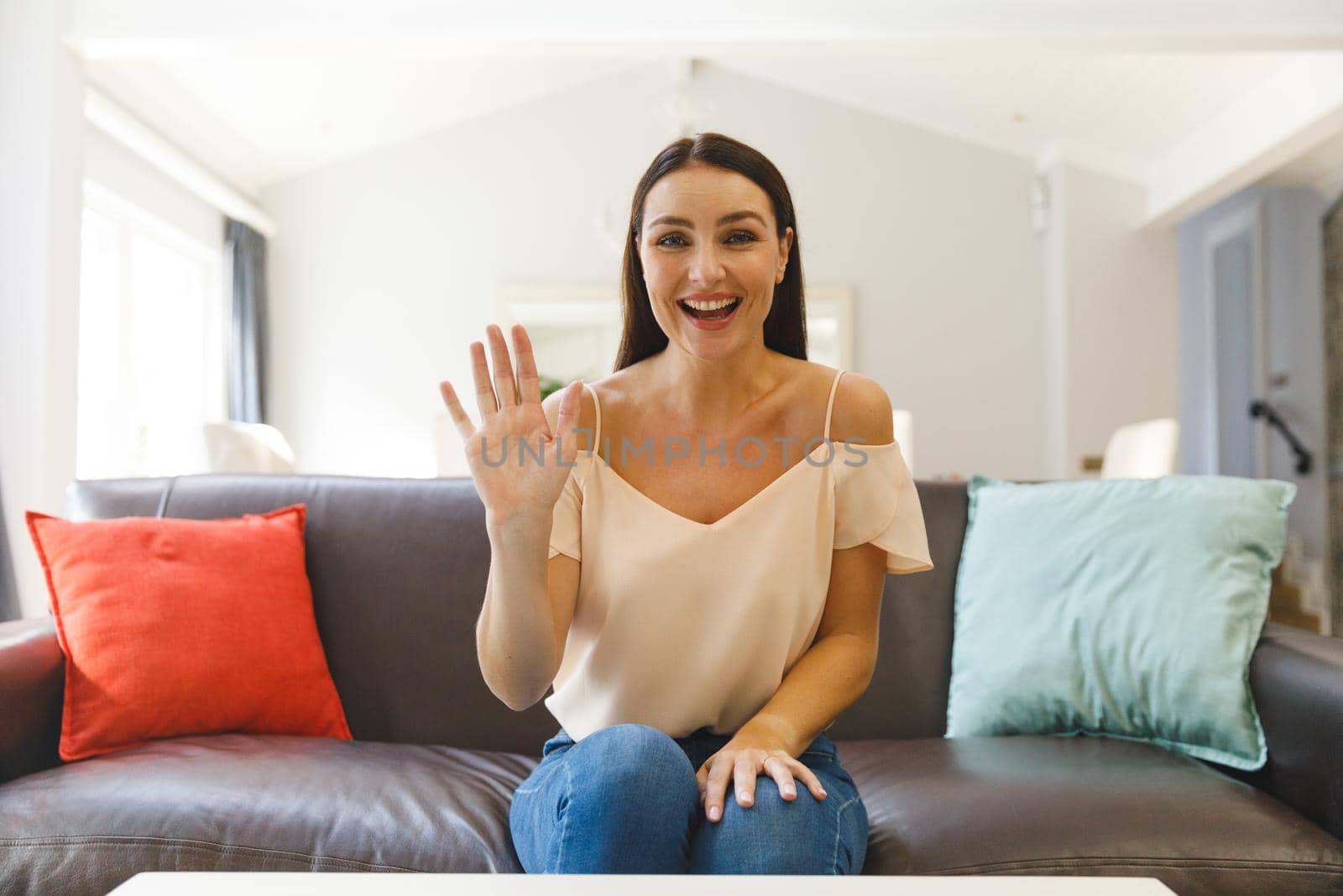 Caucasian woman sitting on couch having video call in living room, smiling and waving. keeping in touch, leisure time at home with communication technology.