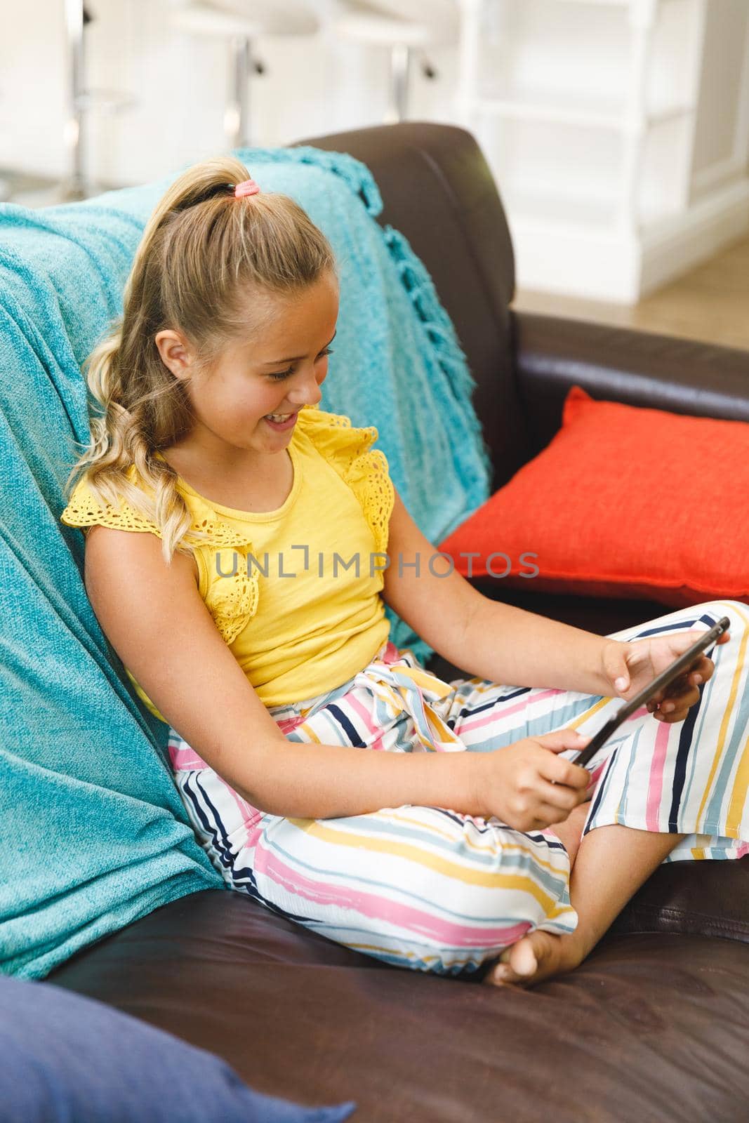 Laughing caucasian girl sitting on couch and using tablet in living room. childhood leisure time, fun and discovery at at home using technology.