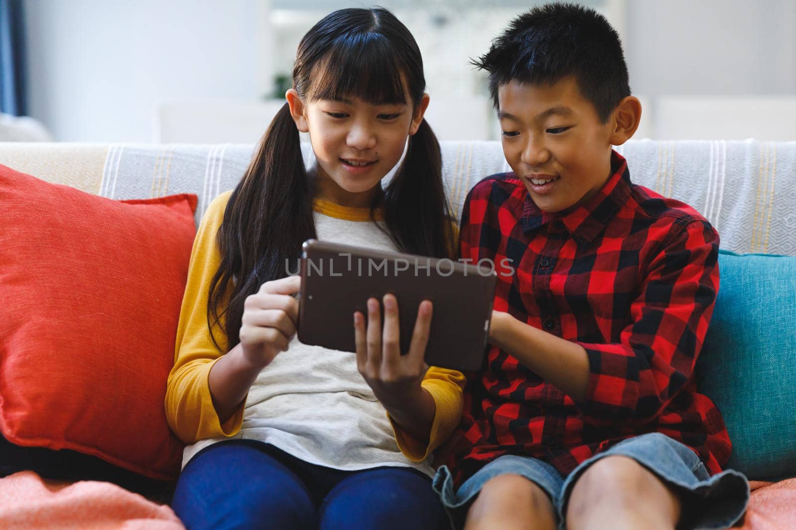 Smiling asian brother and sister sitting on couch and using tablet. childhood leisure time at home with technology.