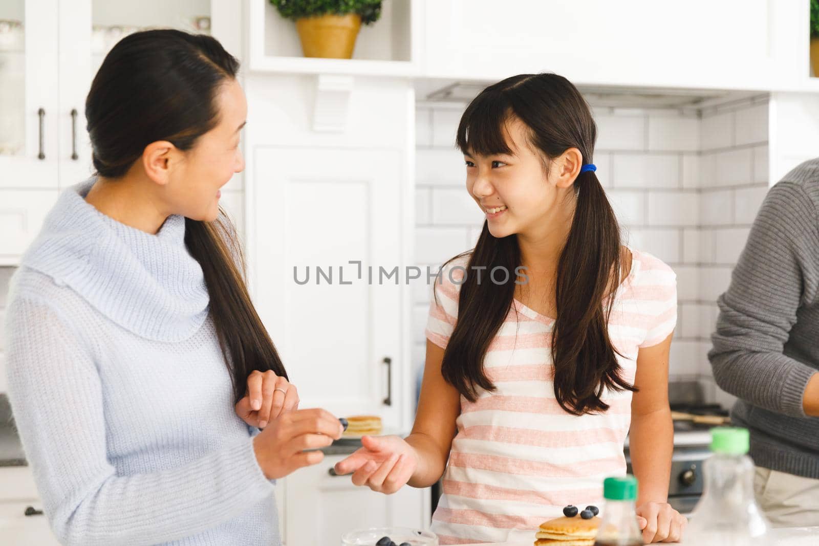 Smiling asian mother and daughter talking while preparing breakfast in kitchen with father. family enjoying preparing meal together at home.