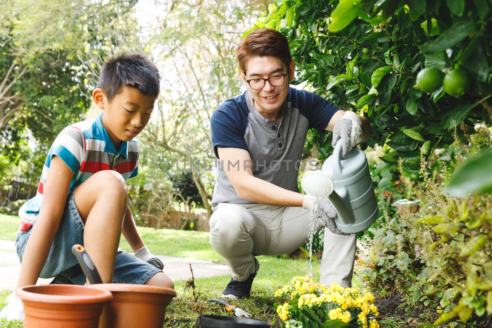 Happy asian father and son smiling, wearing gloves and watering plants together in garden by Wavebreakmedia