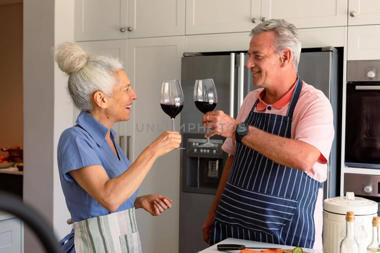 Happy caucasian senior couple standing in kitchen, drinking wine and preparing meal together. healthy retirement lifestyle at home.