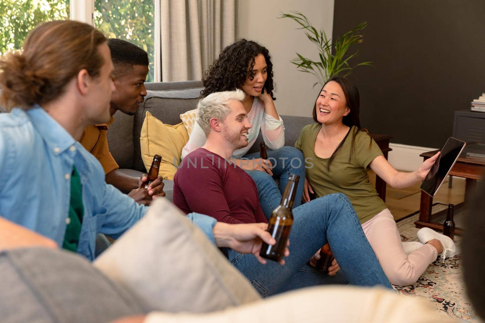 Group of happy diverse female and male friends drinking beer together and using tablet. socialising with friends at home.