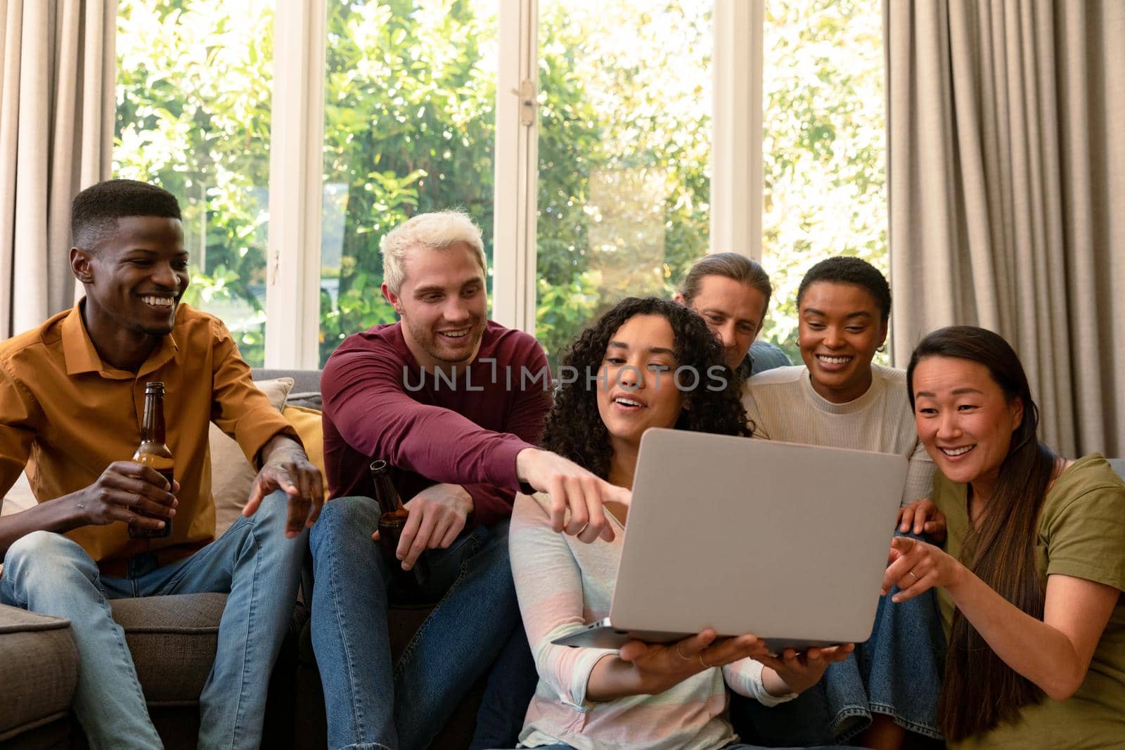 Group of happy diverse female and male friends drinking beer together and using laptop. socialising with friends at home.