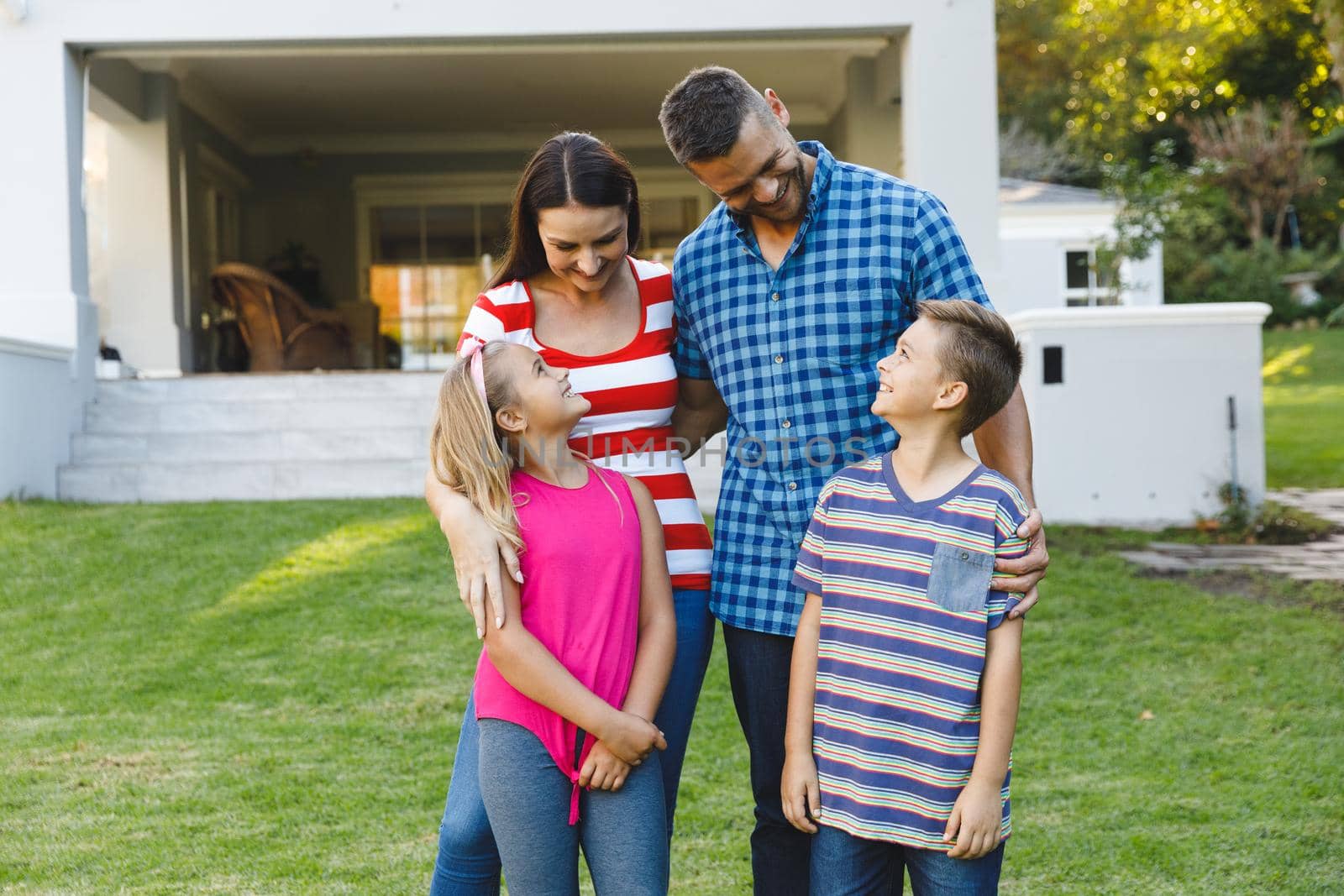 Smiling caucasian parents with son and daughter outside house embracing in garden. family enjoying leisure time together at home.