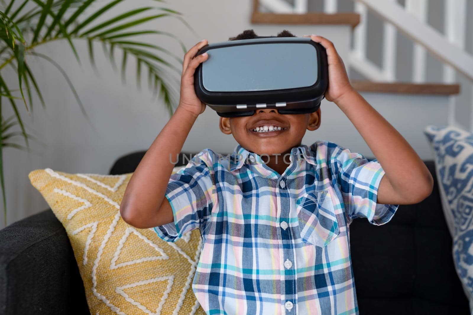 Happy african american boy sitting on sofa, using vr headset and having fun. childhood, leisure and discovery using technology at home.
