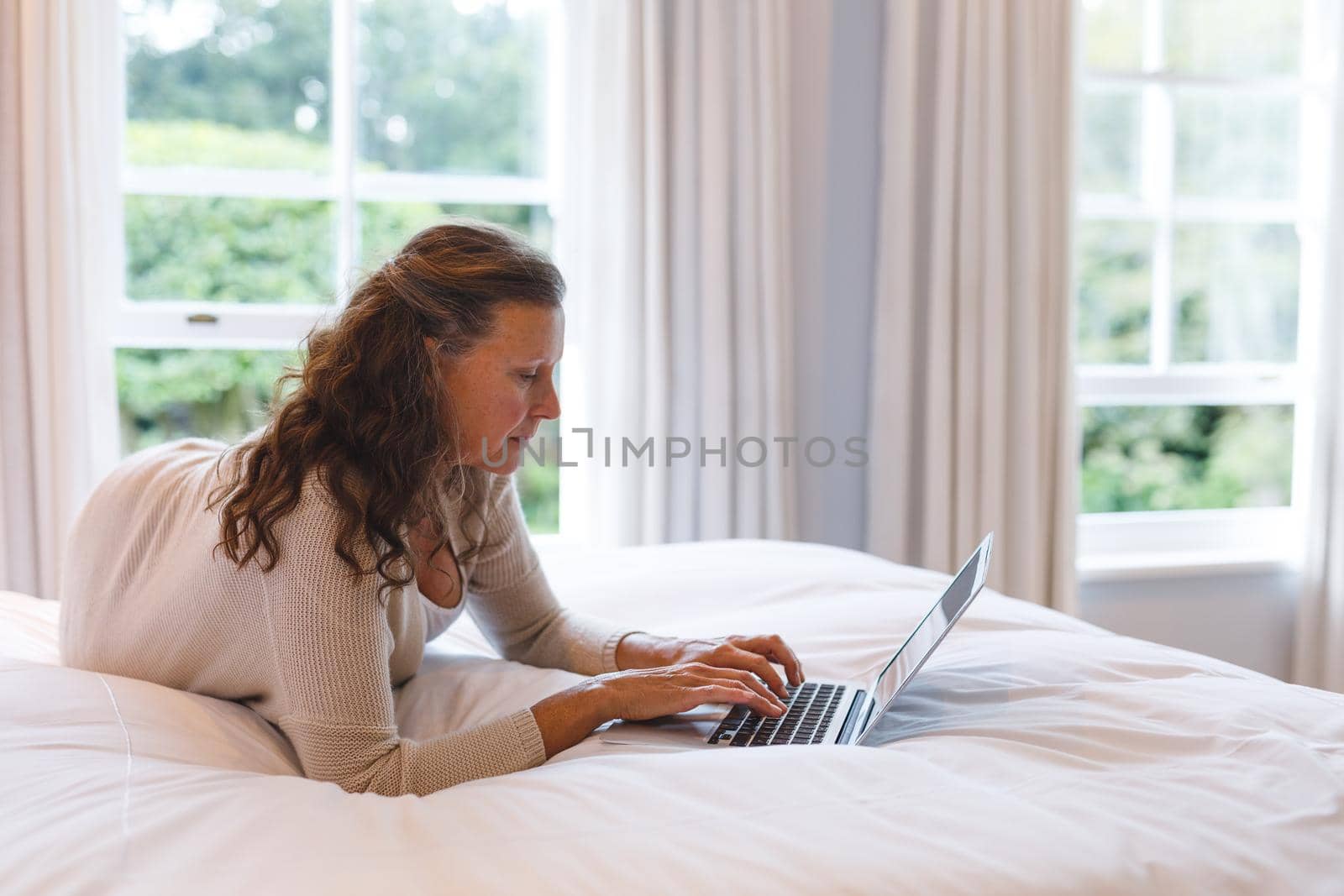 Happy senior caucasian woman in bedroom lying on bed, using laptop. retirement lifestyle, spending time alone at home with technology.