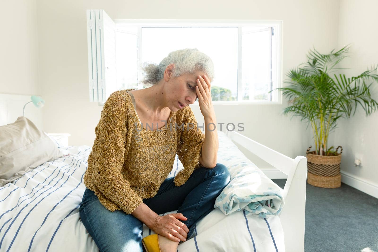 Tired caucasian senior woman with headache sitting on bed at home by Wavebreakmedia
