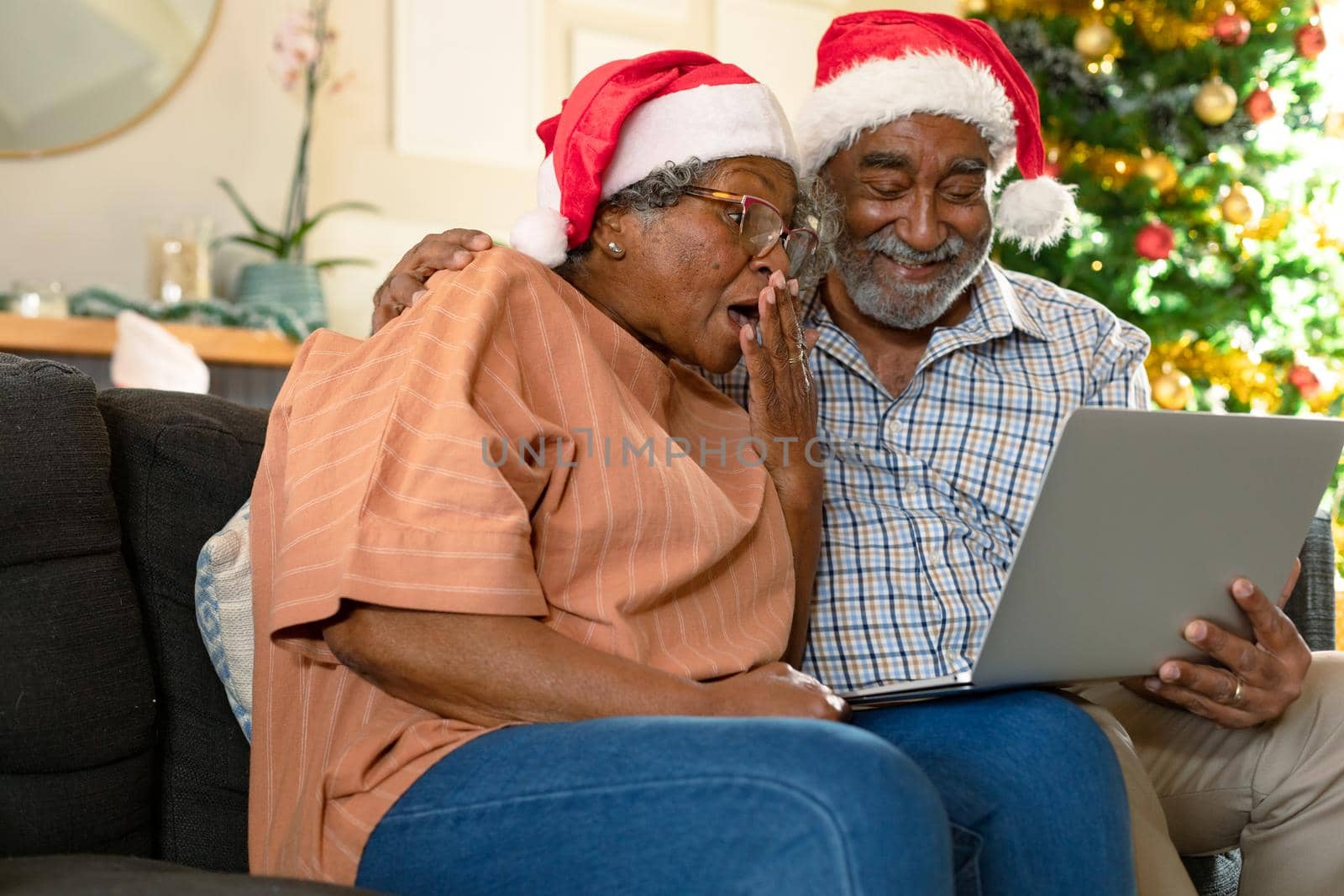 Suprised african american couple wearing santa hats having video call at christmas time. christmas, festivity and communication technology at home.