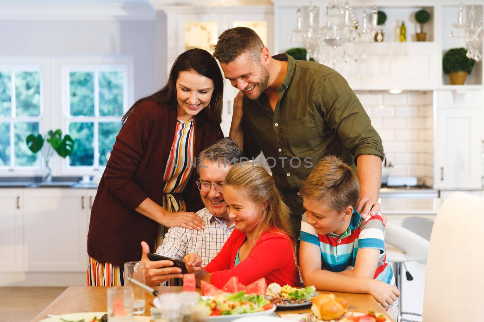 Happy caucasian grandfather and parents with son and daughter looking at smartphone at dinner table. family spending time together at home. able