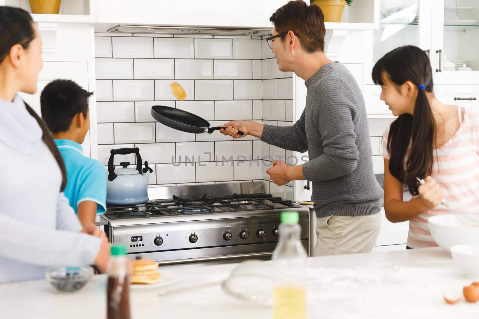 Asian mother, son and daughter watching father tossing pancake for breakfast in kitchen by Wavebreakmedia