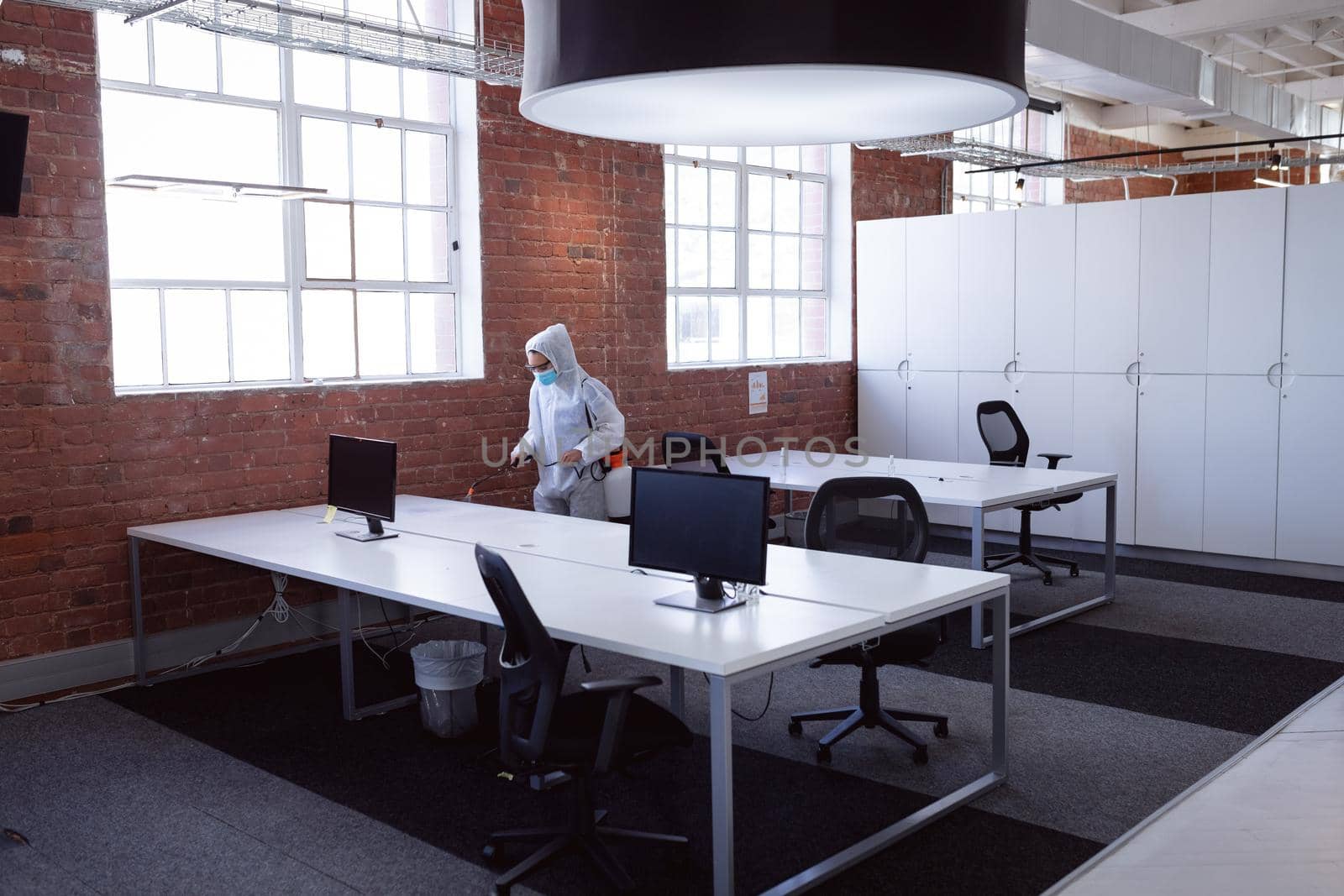 Cleaner wearing ppe suit, glasses and mask disinfecting office workspace. hygiene in business at a modern office during coronavirus covid 19 pandemic.