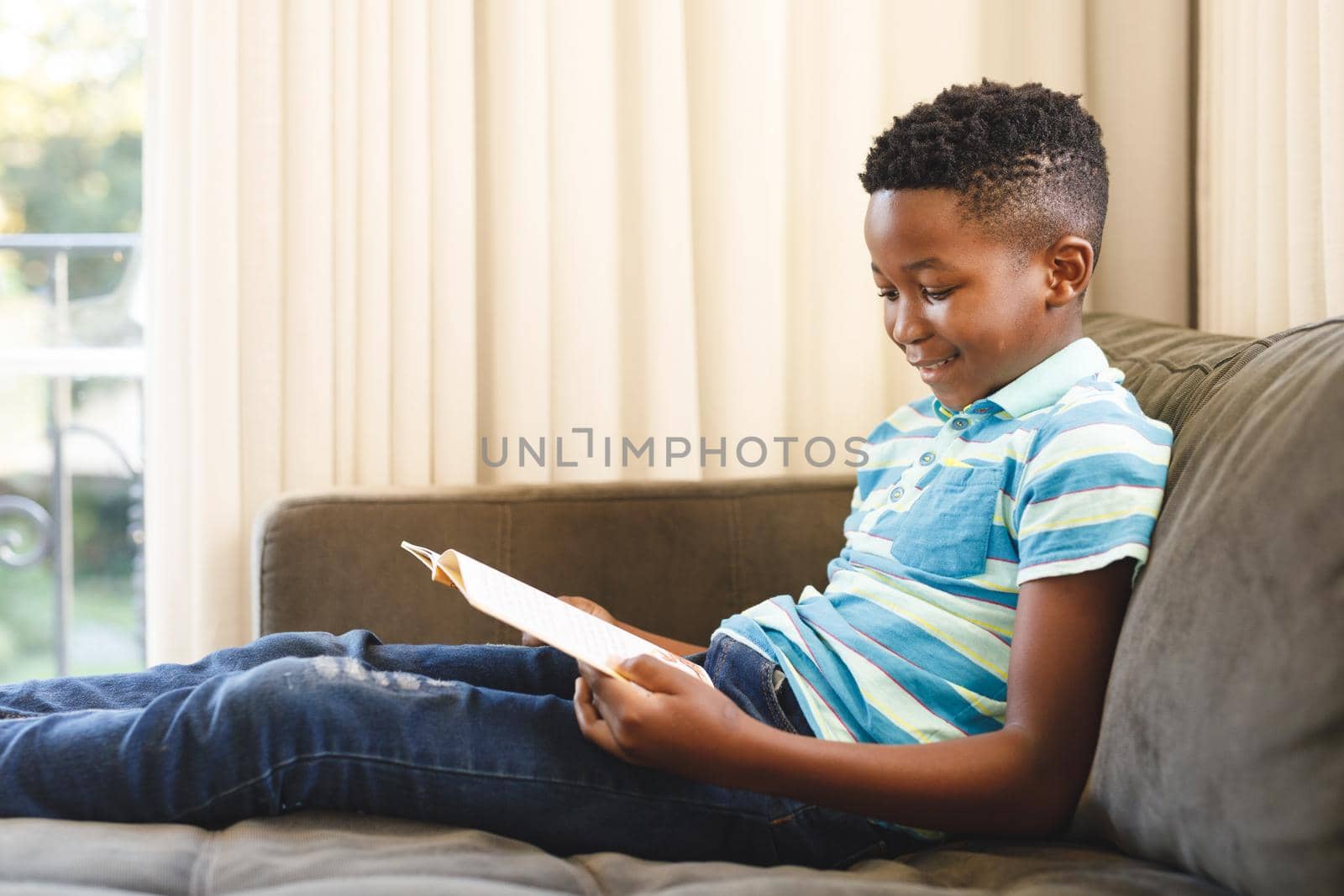 Smiling african american boy reading book and sitting on couch in living room by Wavebreakmedia