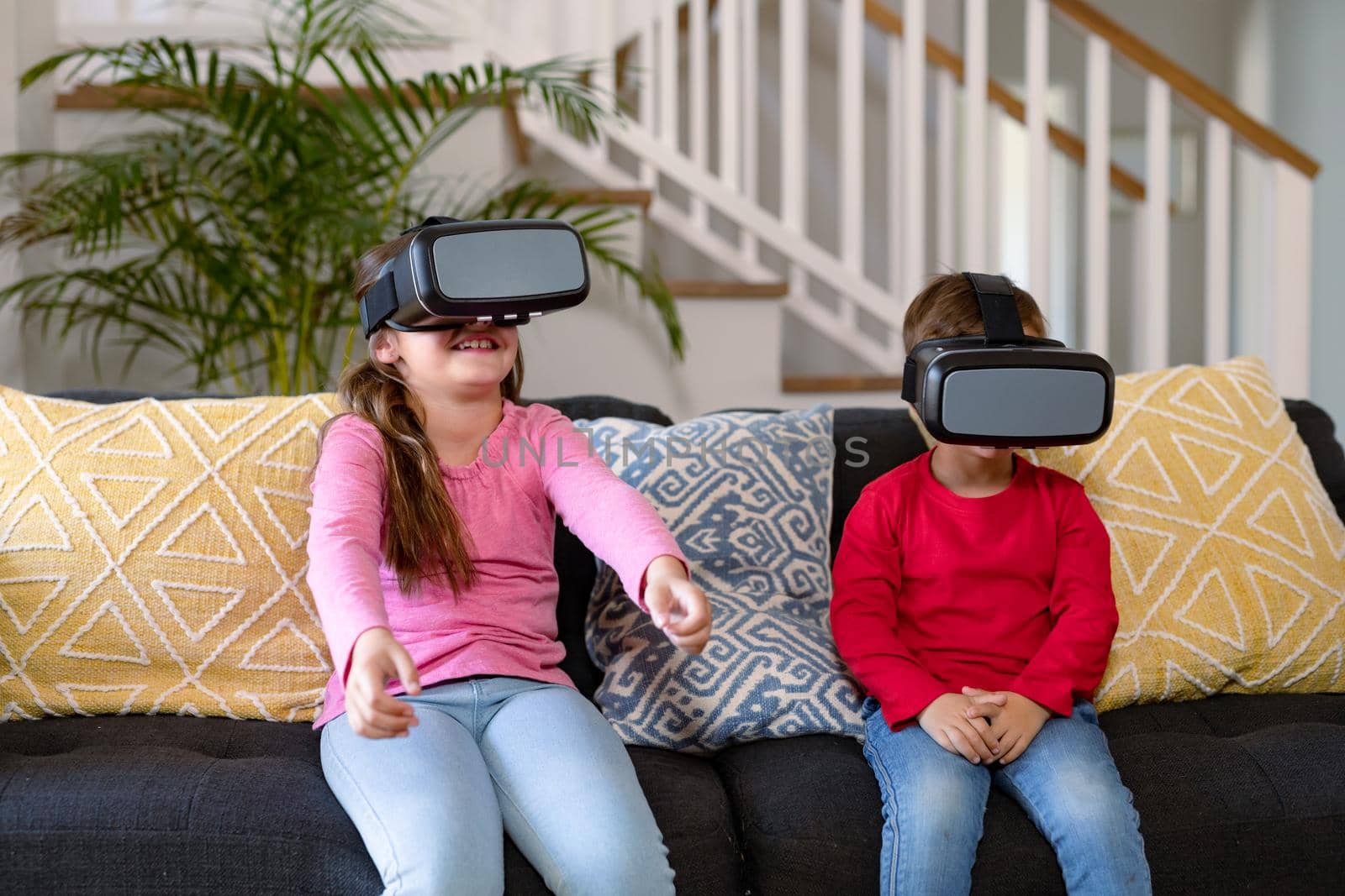 Happy caucasian siblings sitting on sofa, wearing vr headset and having fun. childhood, leisure and discovery using technology at home.