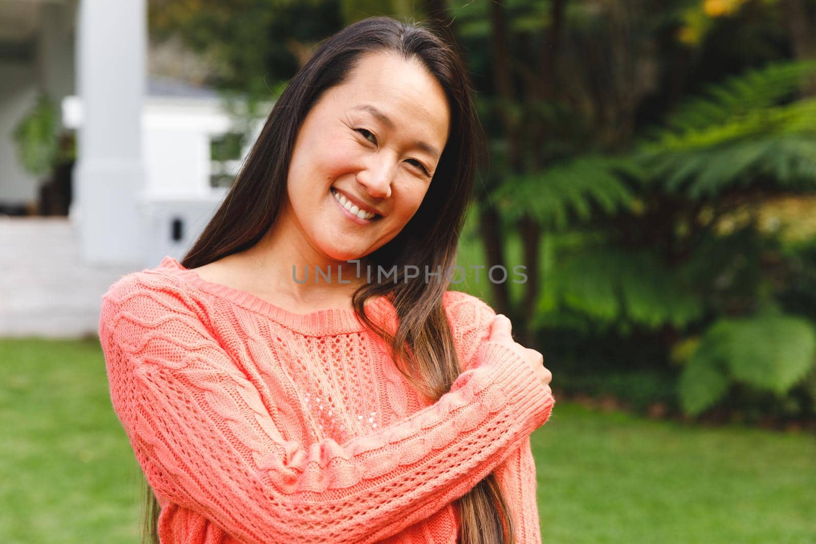 Portrait of happy asian woman smiling in garden outside family home wearing pink sweater. enjoying leisure time alone at home.