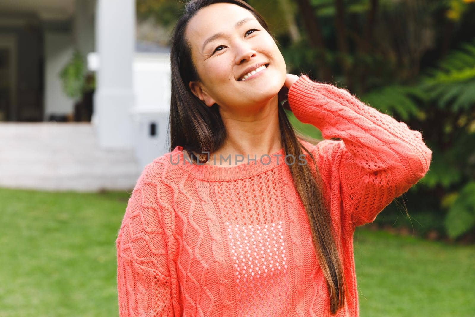 Portrait of happy asian woman smiling in garden outside family home wearing pink sweater. enjoying leisure time alone at home.