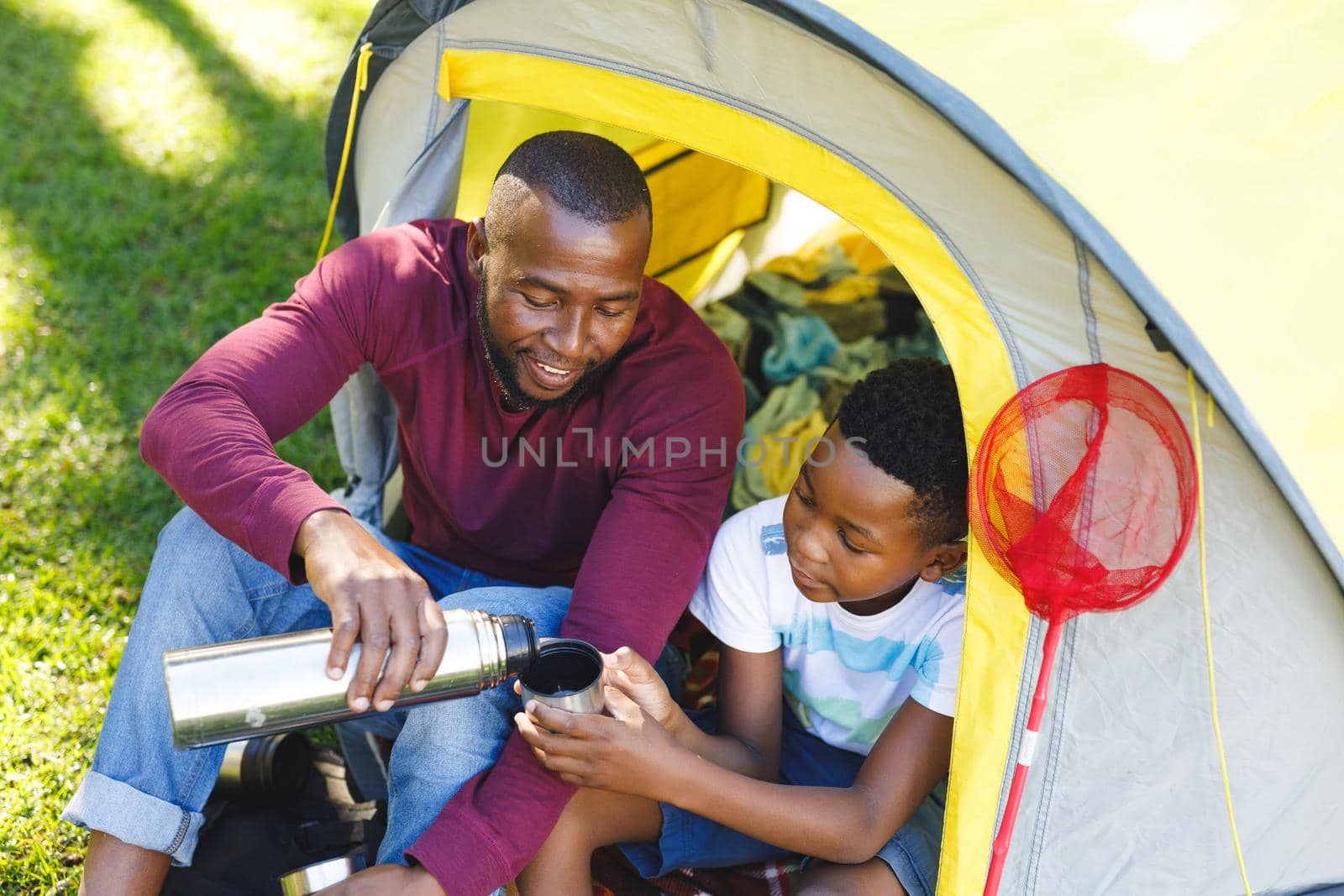 African american father with son having fun and sitting in tent pouring drinks in garden. family spending time at home.