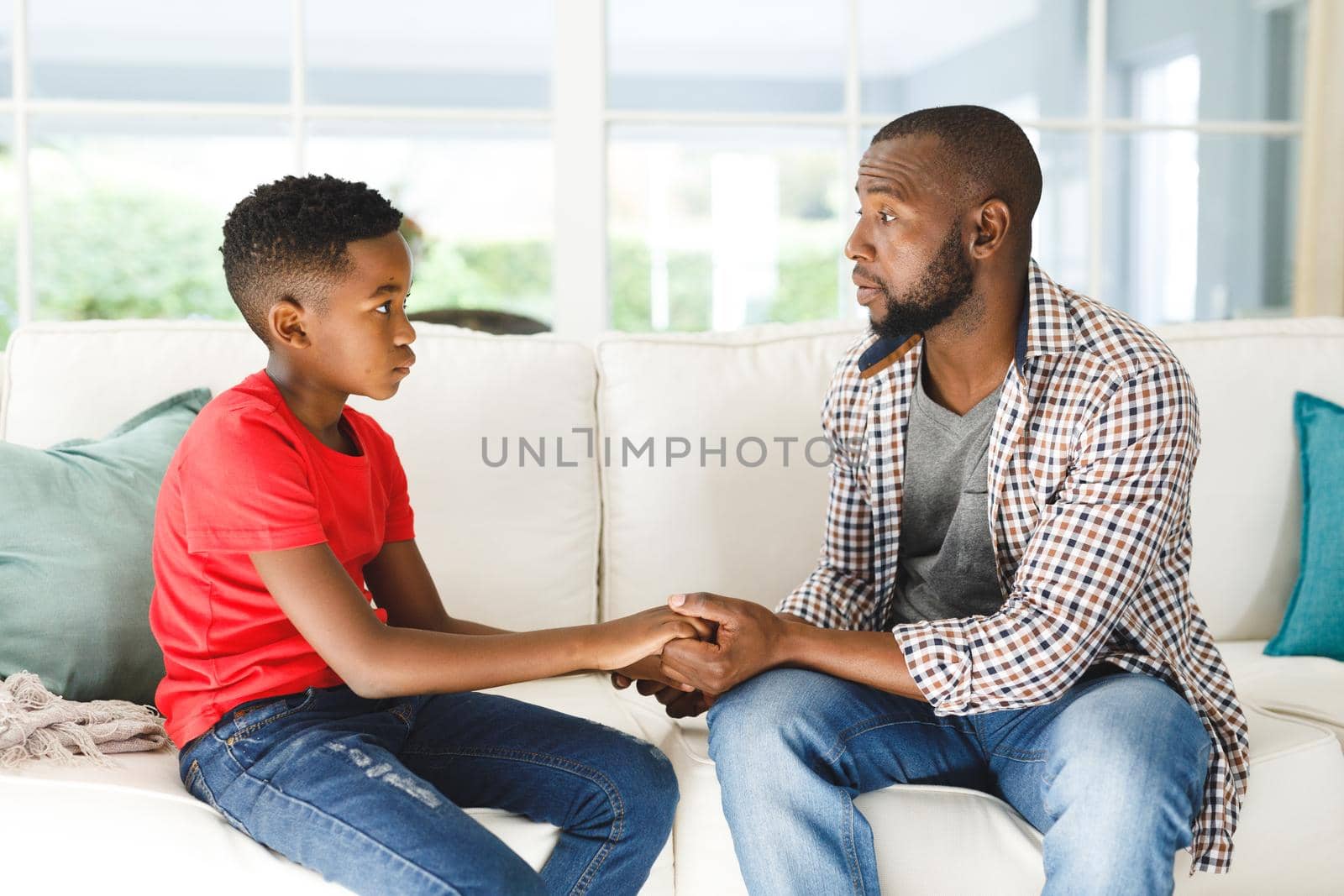 Serious african american father and son sitting on couch in living room talking and holding hands by Wavebreakmedia