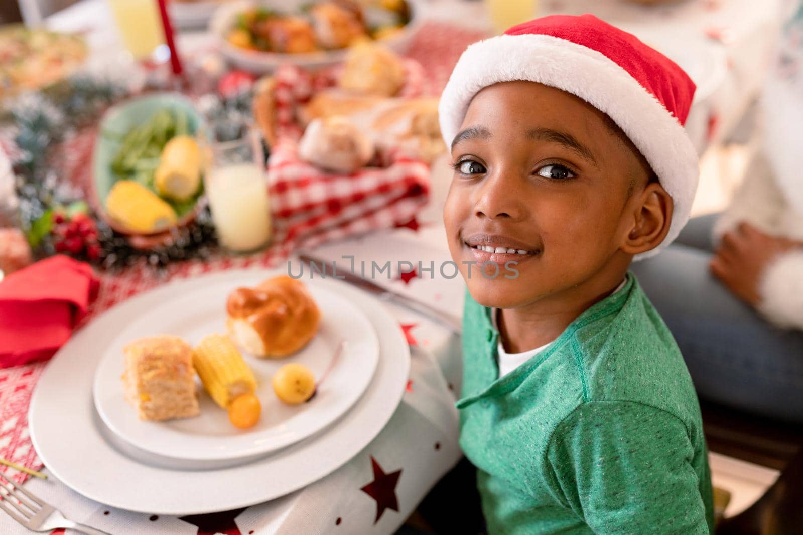 Happy african american boy wearing santa hat, sitting at table, looking at camera. family christmas time and festivity together at home.