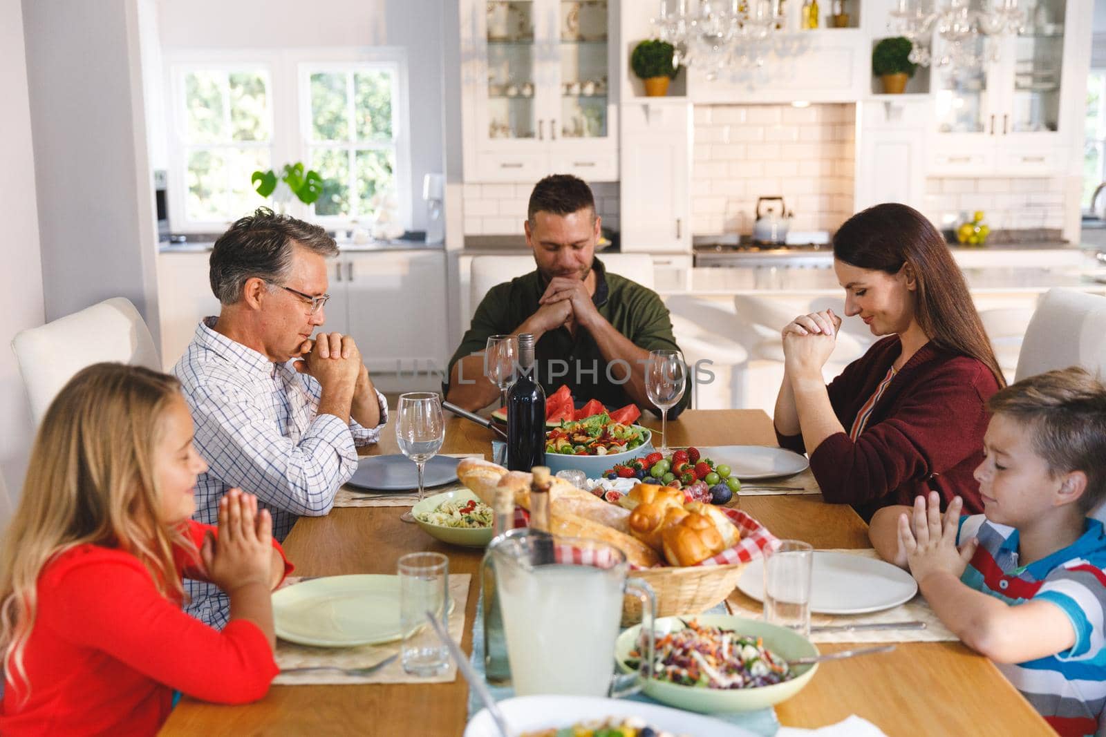 Caucasian grandfather and parents with son and daughter sitting at table and praying before dinner. family enjoying traditional mealtime together at home.