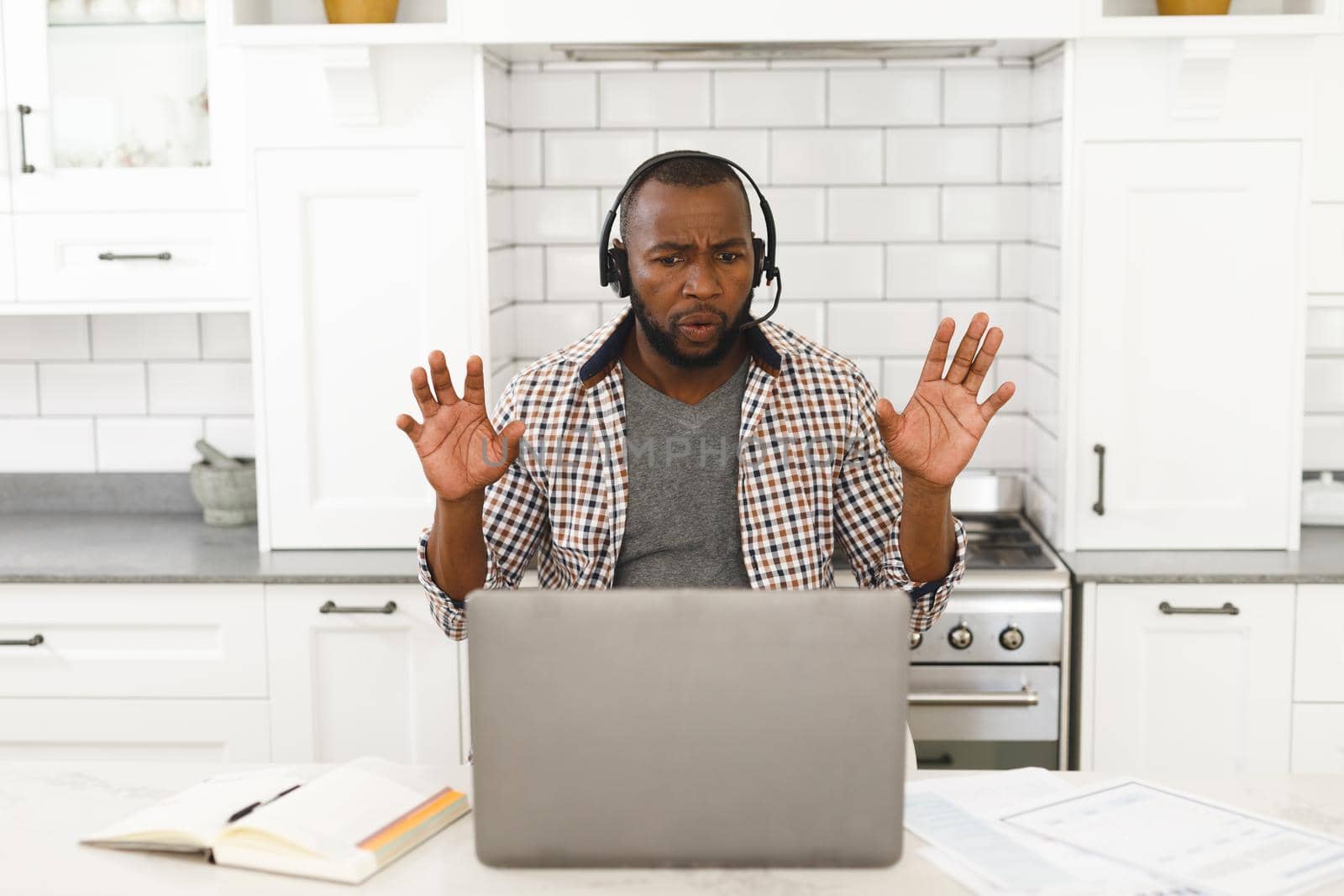 Gesturing african american man sitting in kitchen making video call using laptop and headset by Wavebreakmedia