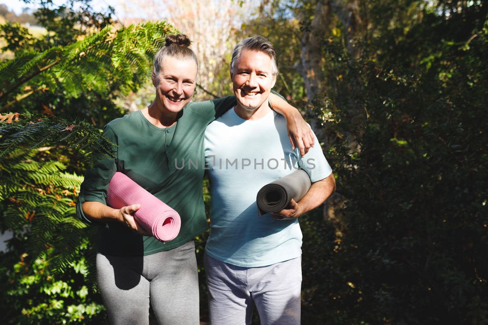 Portrait of happy senior caucasian couple holding yoga mats, looking to camera in sunny garden. healthy retirement lifestyle, spending time at home.