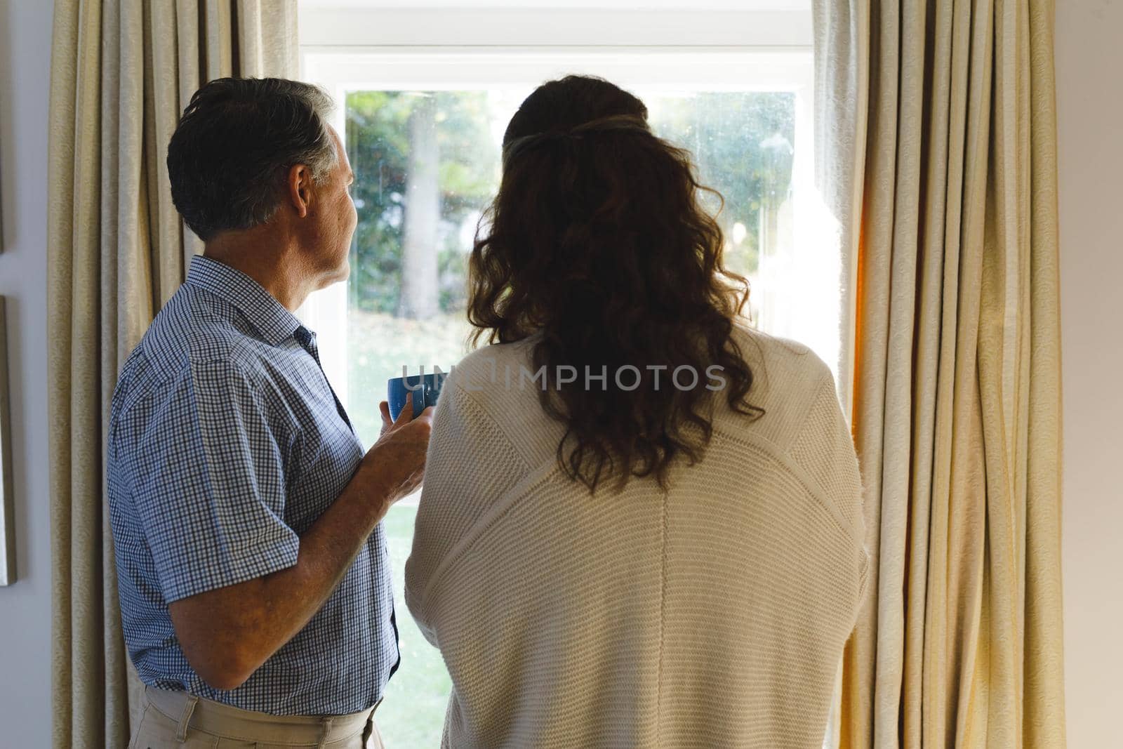 Happy senior caucasian couple standing next to window, holding cups. retirement lifestyle, spending time at home.